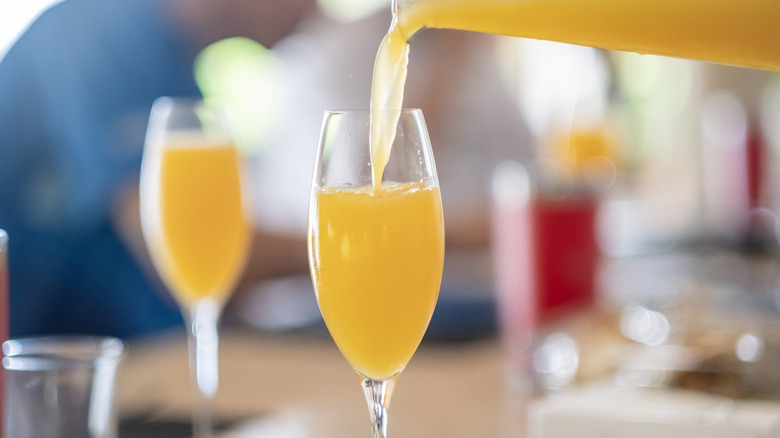Una botella que vierte mimosa en una copa de champán con una copa llena al fondo.