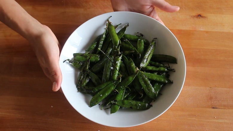 Dos manos sosteniendo un plato blanco de guisantes asados, todavía en sus vainas