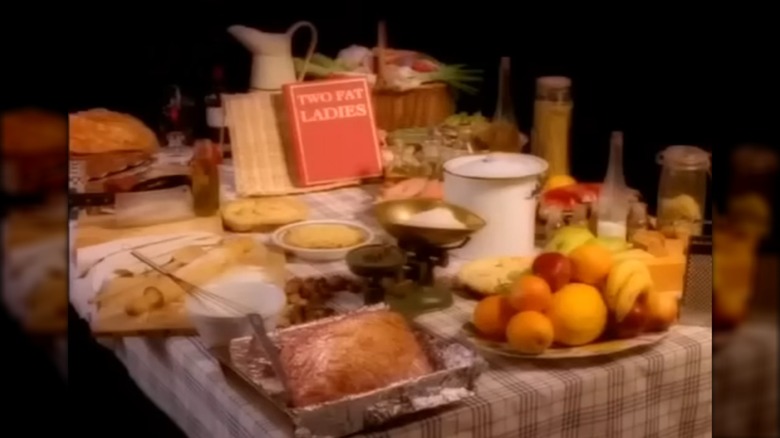 Tarjeta de título de Two Fat Ladies con el nombre del programa en un libro rojo, sobre una mesa cubierta de comida.
