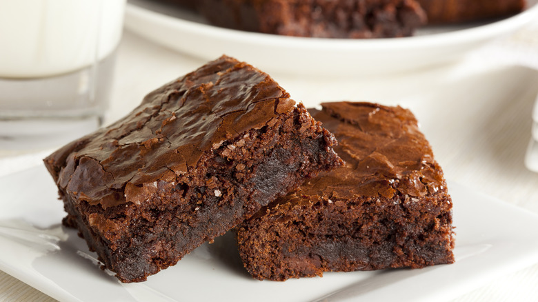 Dos rebanadas de brownies de chocolate en un plato blanco con el borde de un plato lleno de brownies al fondo.