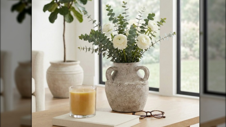 Un jarrón blanquecino con flores blancas junto a vasos rojos y una vela.