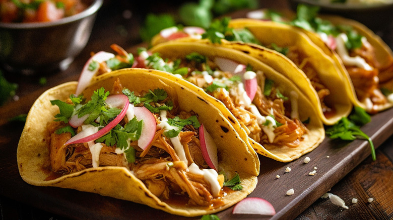 Una fila de tacos de pollo sobre una tabla adornados con cilantro y rodajas de rábano.