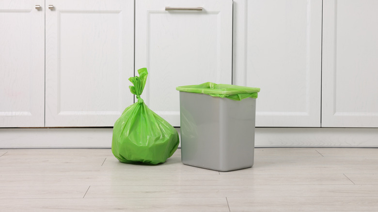 Bolsa de basura verde llena que ha sido atada junto al contenedor con una bolsa nueva en una cocina con gabinetes blancos.