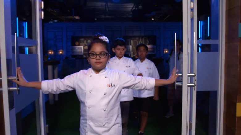 4 niños pequeños con chaquetas blancas de chef que dicen "TCJR" caminando dramáticamente a través de una puerta
