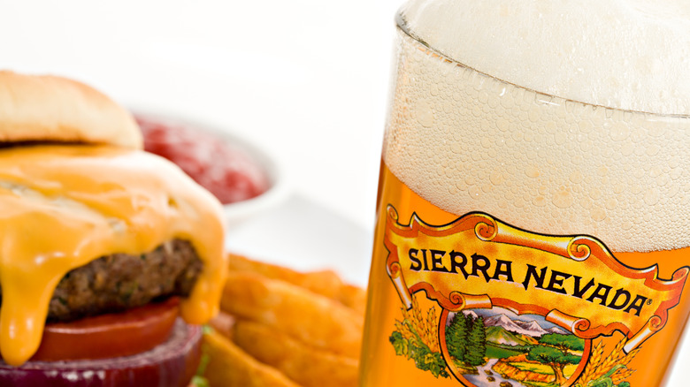 Logotipo de Sierra Nevada Brewing Co. en un vaso de cerveza lleno de cerveza dorada, con una hamburguesa con queso y papas fritas al lado