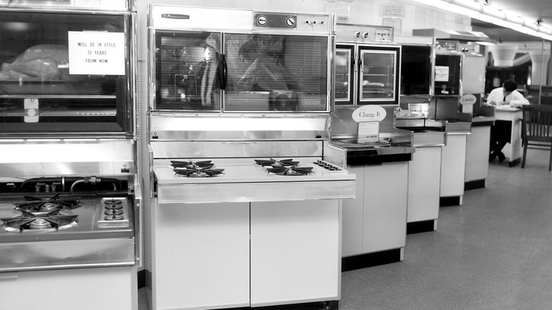 Imagen en blanco y negro de una sala de exposición de electrodomésticos de cocina antiguos de los años 60 con hornos y estufas.