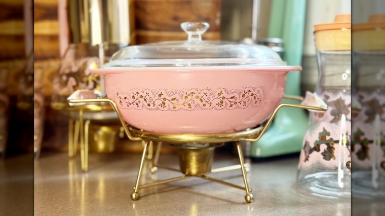 La cacerola Pink Pyrex Duchess con tapa transparente colocada sobre un soporte calentador dorado sobre una encimera