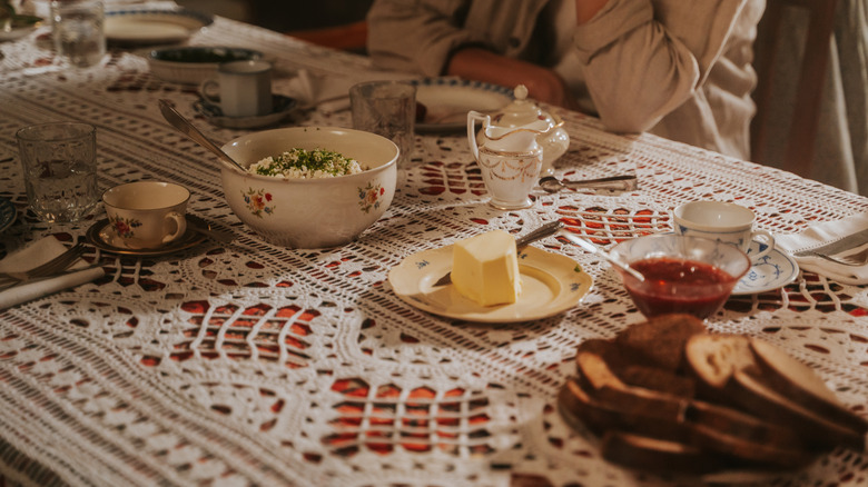 Persona sentada a la mesa del comedor con mantel de encaje, además de pan y mantequilla, platos.