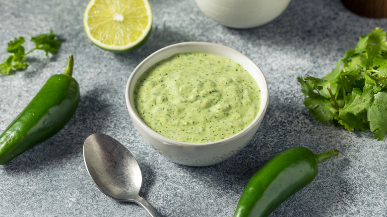 Salsa verde peruana en un recipiente blanco rodeada de cilantro, limas y jalapeños