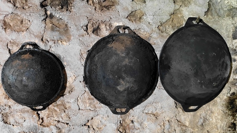Tres woks de hierro fundido de diferentes tamaños con fondos redondos colgados en una pared de piedra