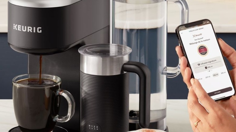 Cafetera Keurig sirviendo café en una taza con la mano sosteniendo el teléfono con la aplicación Keurig abierta