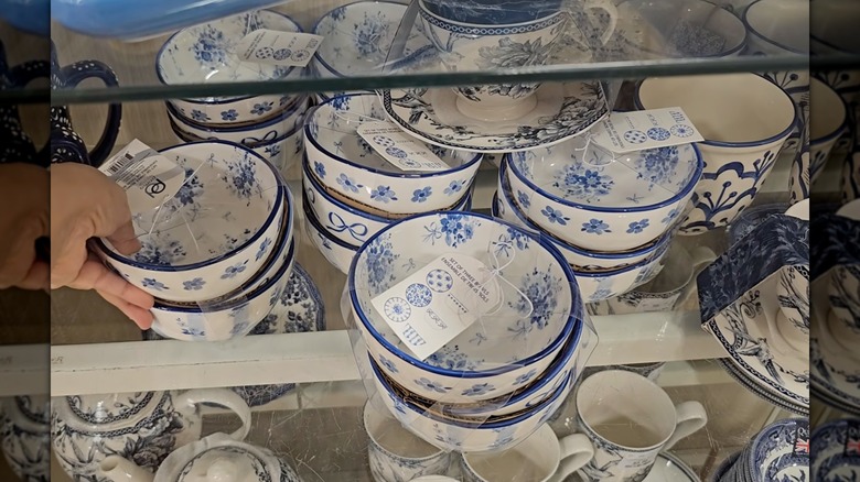 Una mano tocando un juego de tazones azules y blancos, junto a muchos otros juegos de tazones azules y blancos en un estante de HomeGoods
