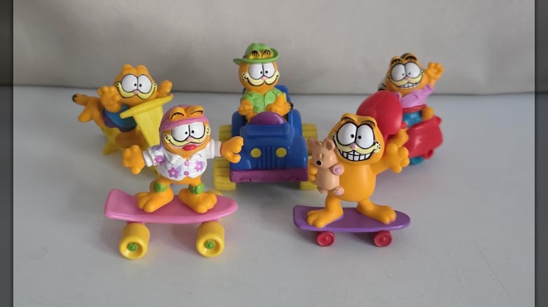 Varias figuras de Garfield en patinetas, scooters y automóviles sobre un fondo blanco.