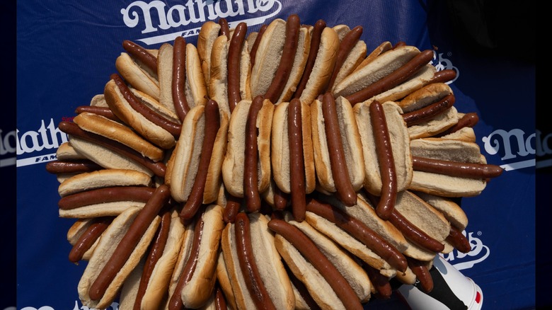 Un montón de hot dogs simples con panecillos en un telón de fondo de Nathan's Famous
