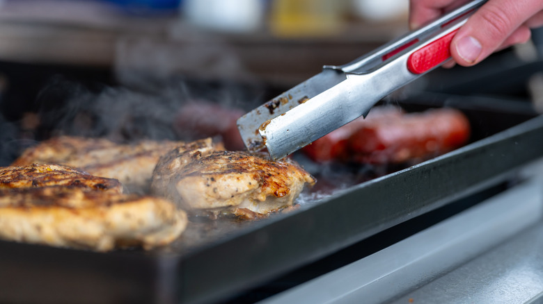 Una mano sosteniendo unas pinzas y volteando pechugas de pollo en una plancha.
