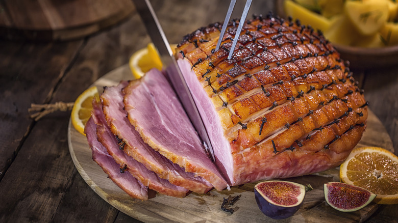 Alguien tallando un jamón cocido con miel con higos y rodajas de limón al lado sobre una tabla de talla de madera