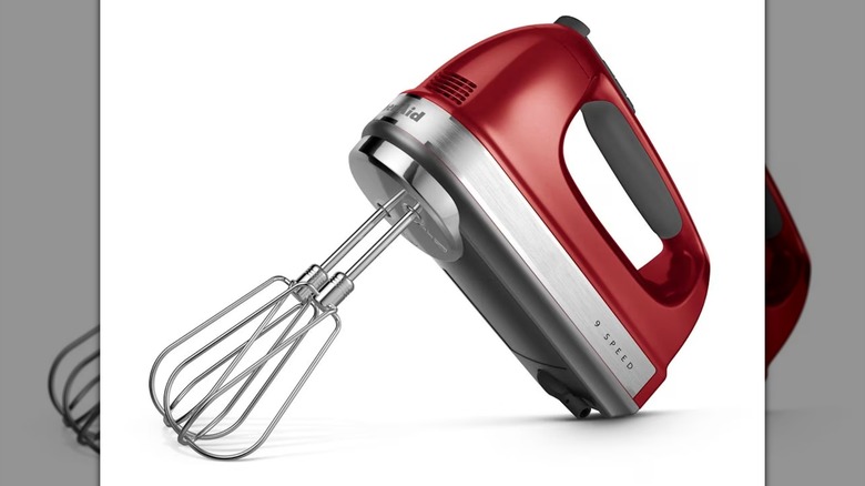 Una batidora de mano KitchenAid de 9 velocidades roja con accesorios para batir sobre un fondo blanco